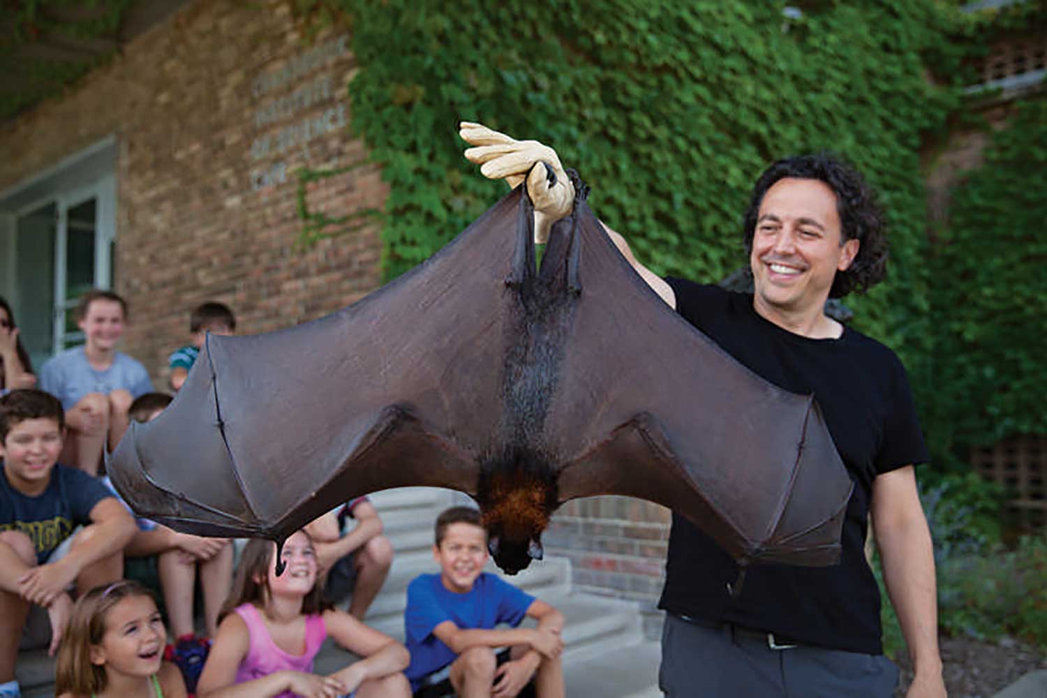 A man holding a very large bat hanging by it's feet in front of a group of delighted children