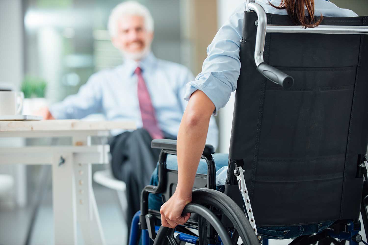 A person in a wheelchair is seen from behind, from the shoulders down. They are sitting across from a businessman, out of focus.