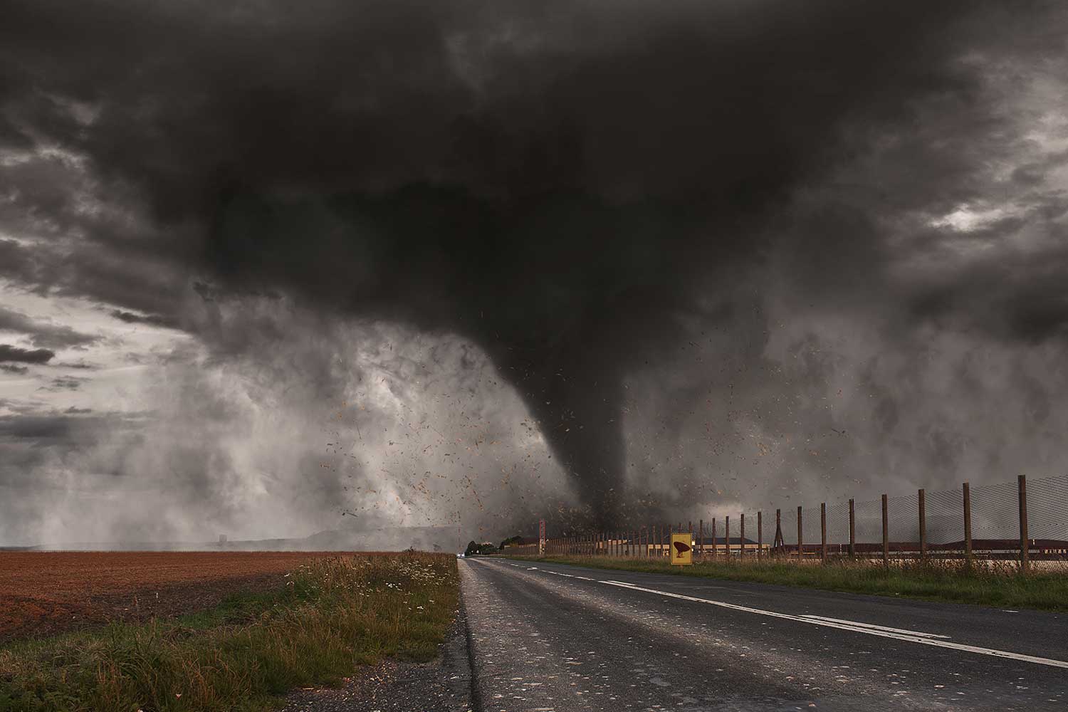 A large tornado is shown approaching a fence alongside a roadway.