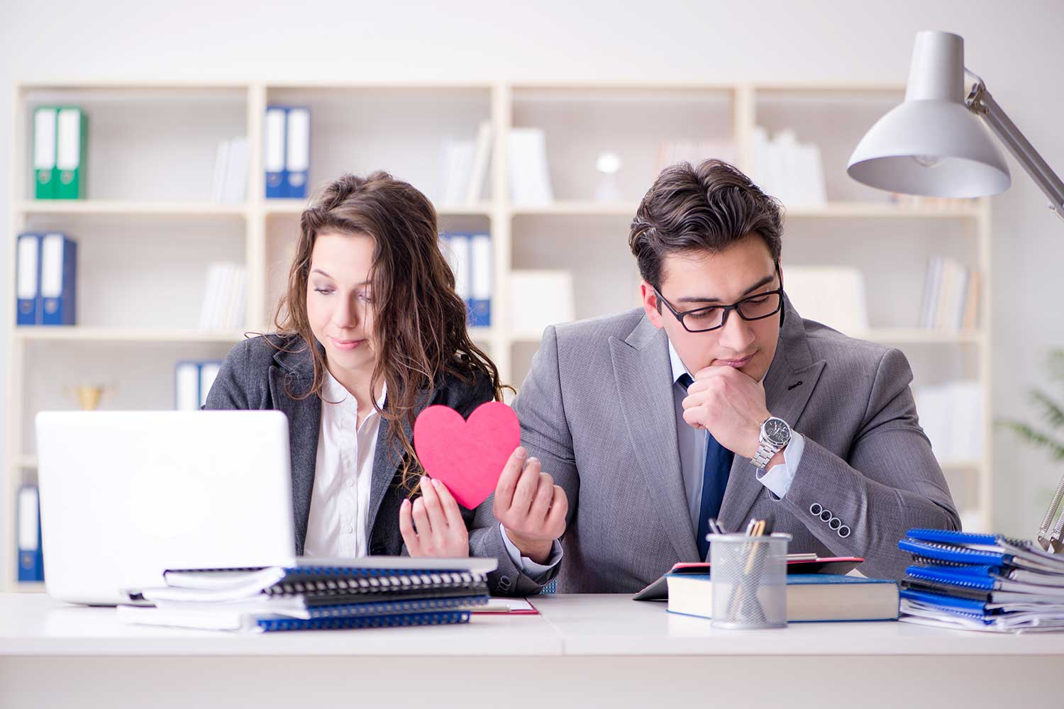 A man and a woman are shown seated, one looking at a laptop, the other at a sheet of paper. They are holding a red paper heart.