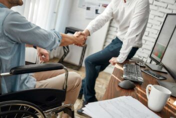 person in wheelchair shaking hands with a person leaning against a desk