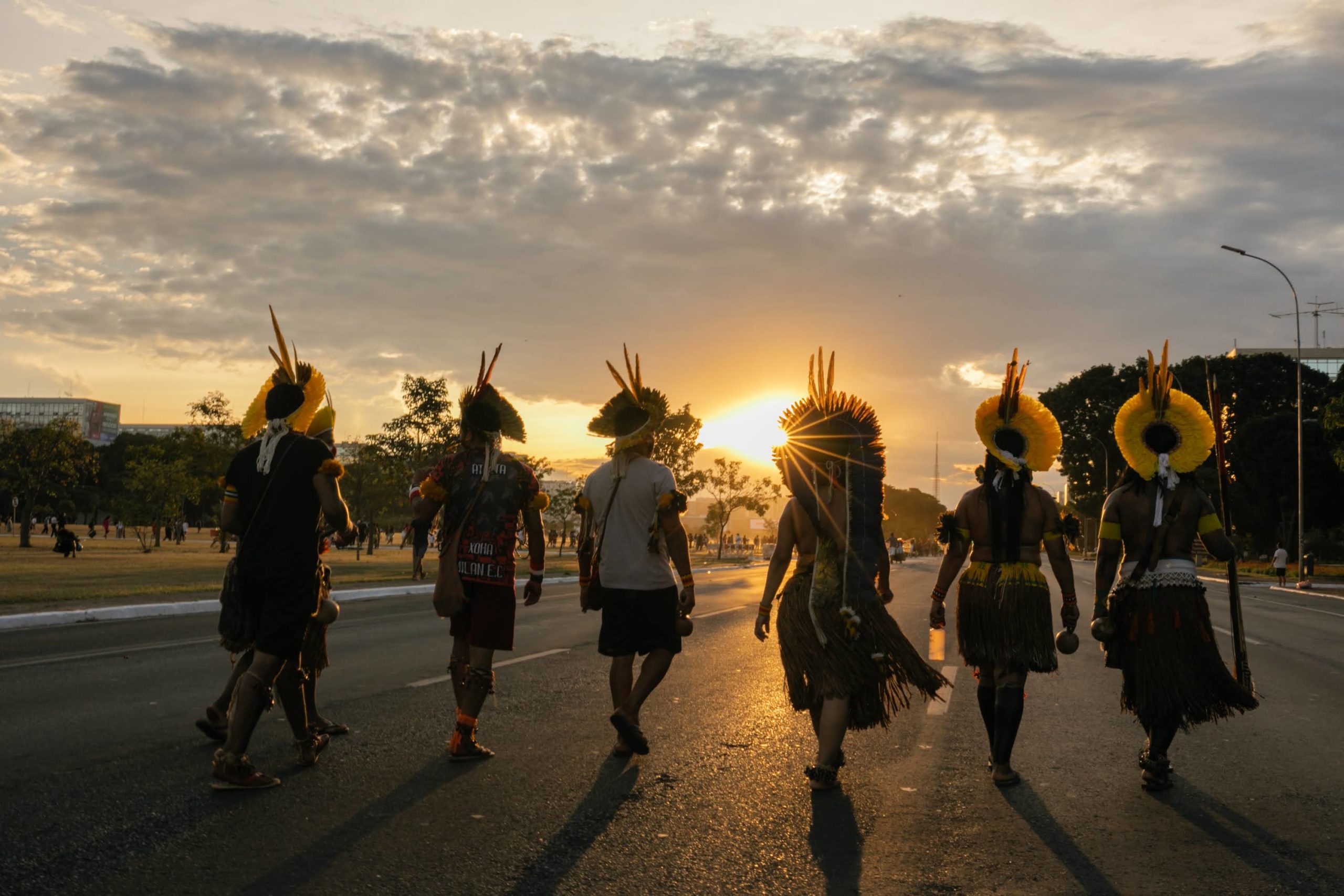 men in native costumes viewed from behind as they walk on a paved street toward the setting sun