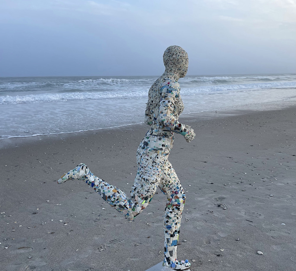 a sculpture of a woman running on the beach made from pieces of plastic found on the beach, with sand and waves in the background