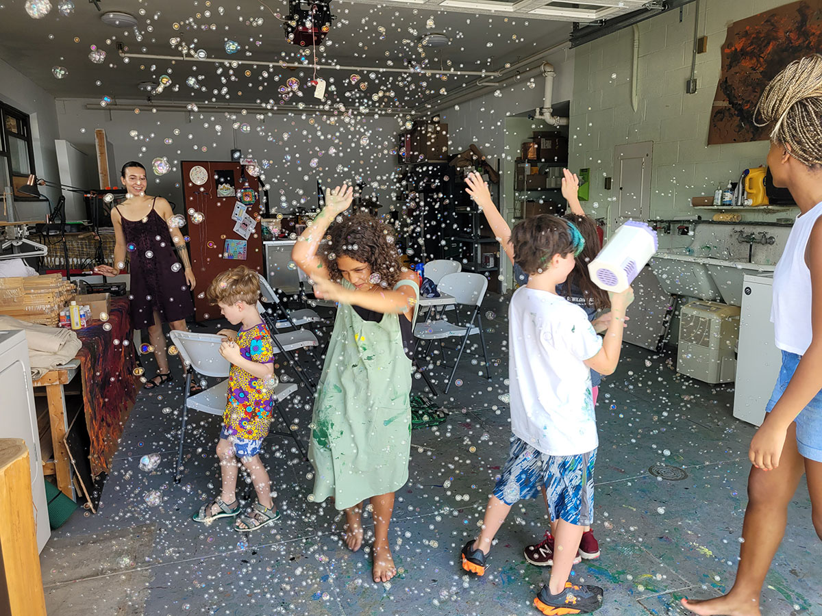 several children dance and play in the bubbles being produced by a machine held by one child, with a woman in the background