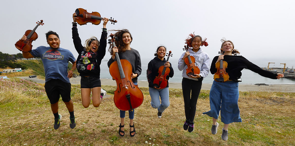 teenage musicians holding their stringed instruments jump for joy in a grassy area near the water's edge