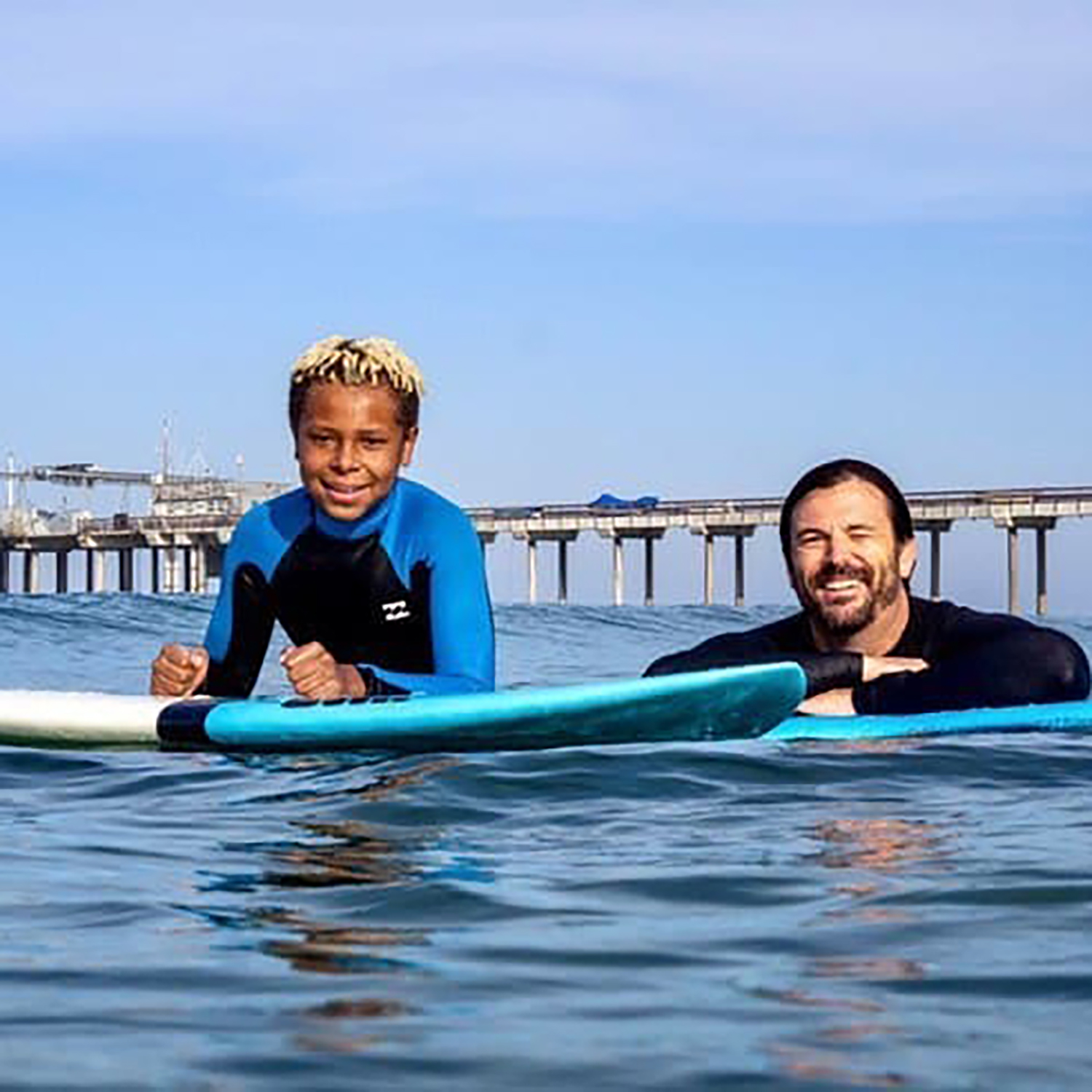 A man and a boy in wetsuits float on a surfboard near a pier