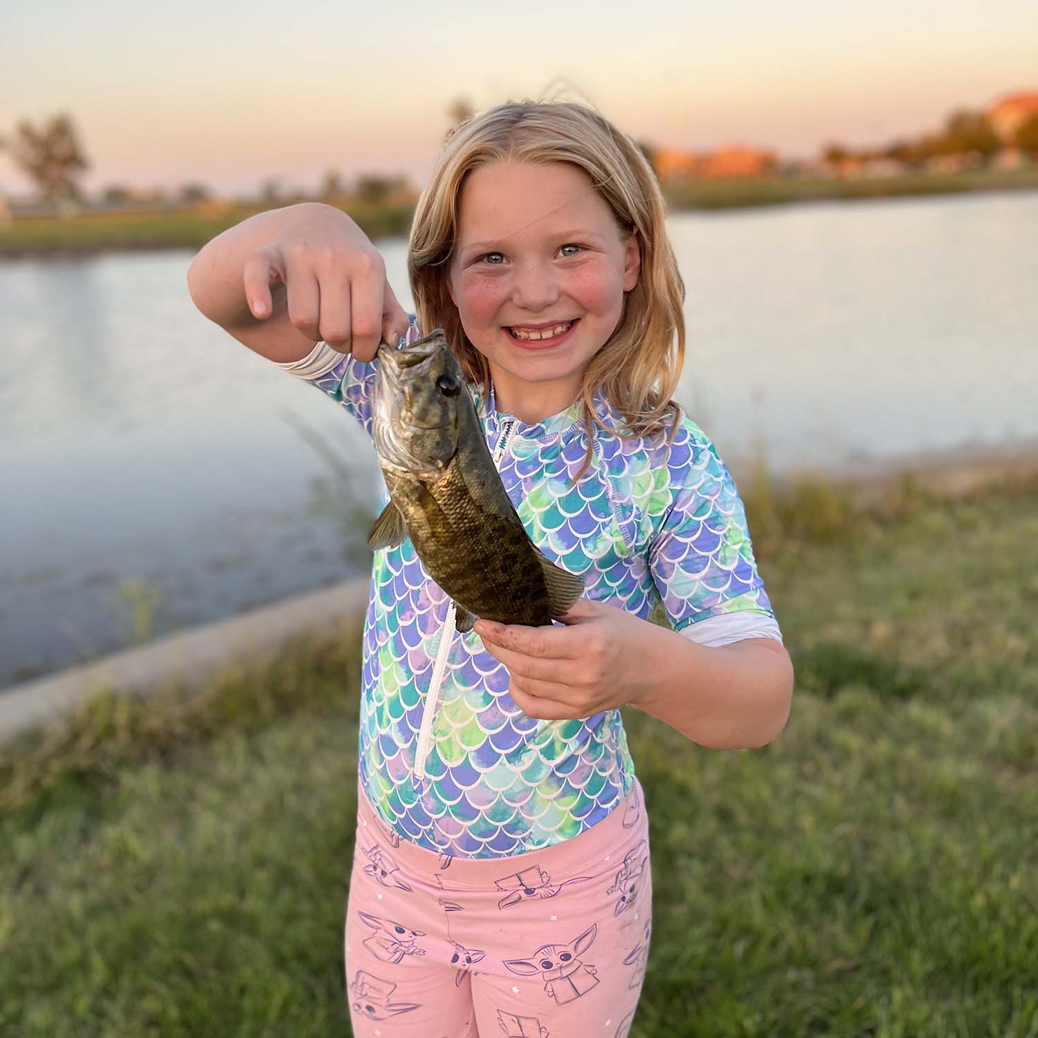 A child proudly holds a small fish she caught with a beautiful lake in the background