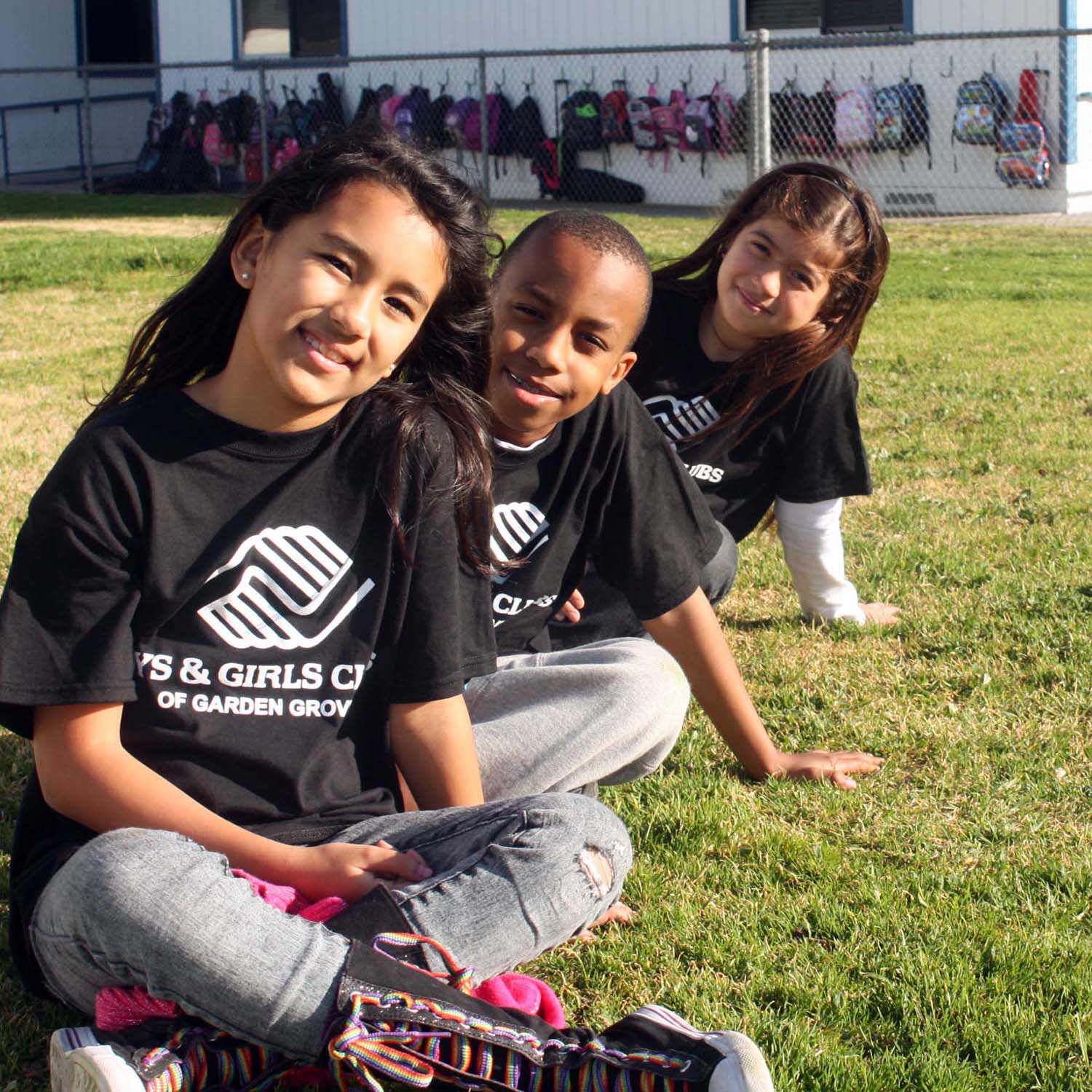 Three children wearing Boys & Girls Club t-shirts sitting on a green sports field