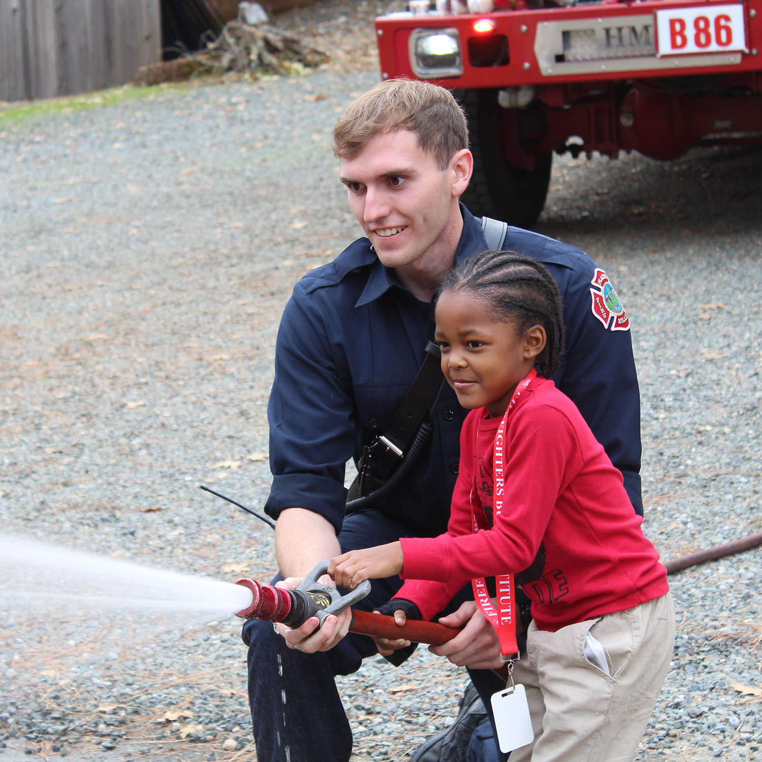 A firefighter helps a young child hold a firehose in front of a fire truck
