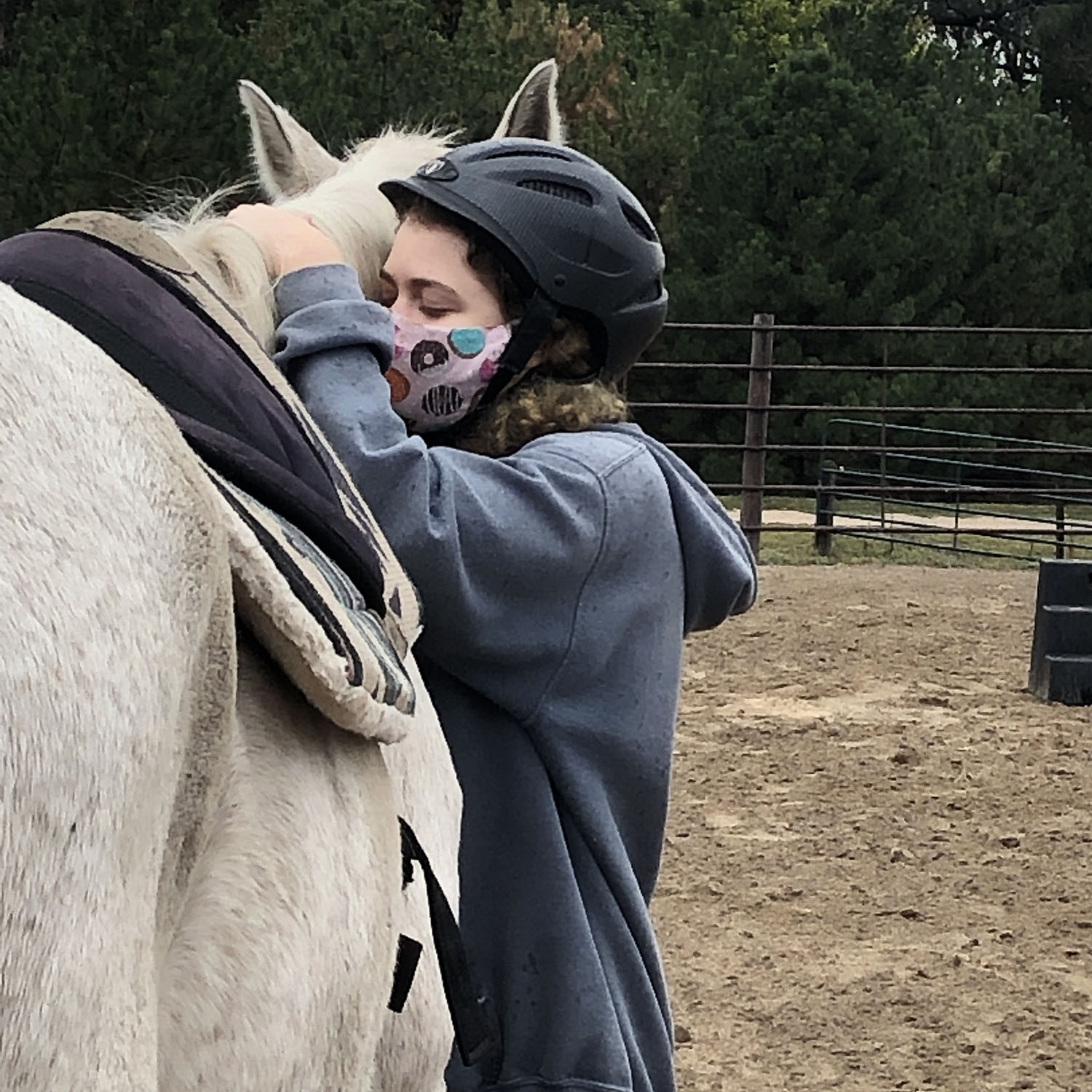 a teen with a facemask hugs a white horse in an outdoor fenced area