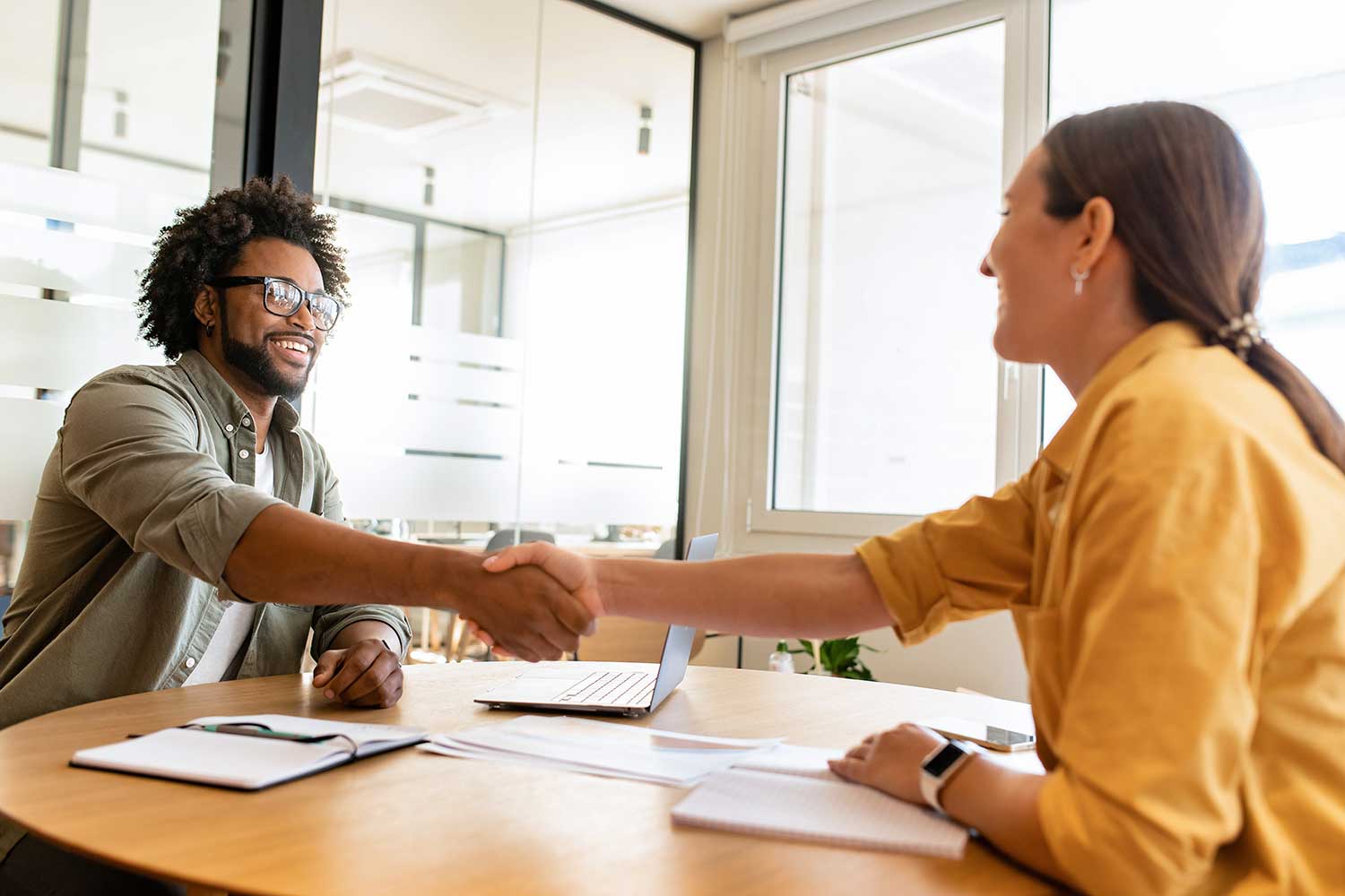 a man and woman smile as they shake hands while sitting at a table with papers and a laptop