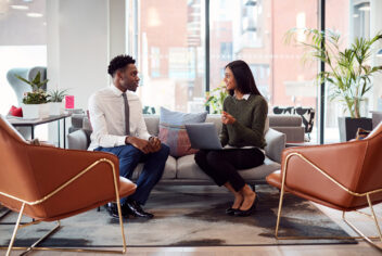two people sitting on a couch in business attire having a conversation while one holds a laptop