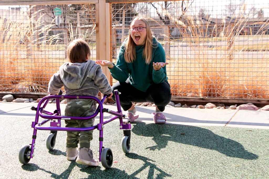 a young woman squatting at a child's level encourages him to walk using a walker