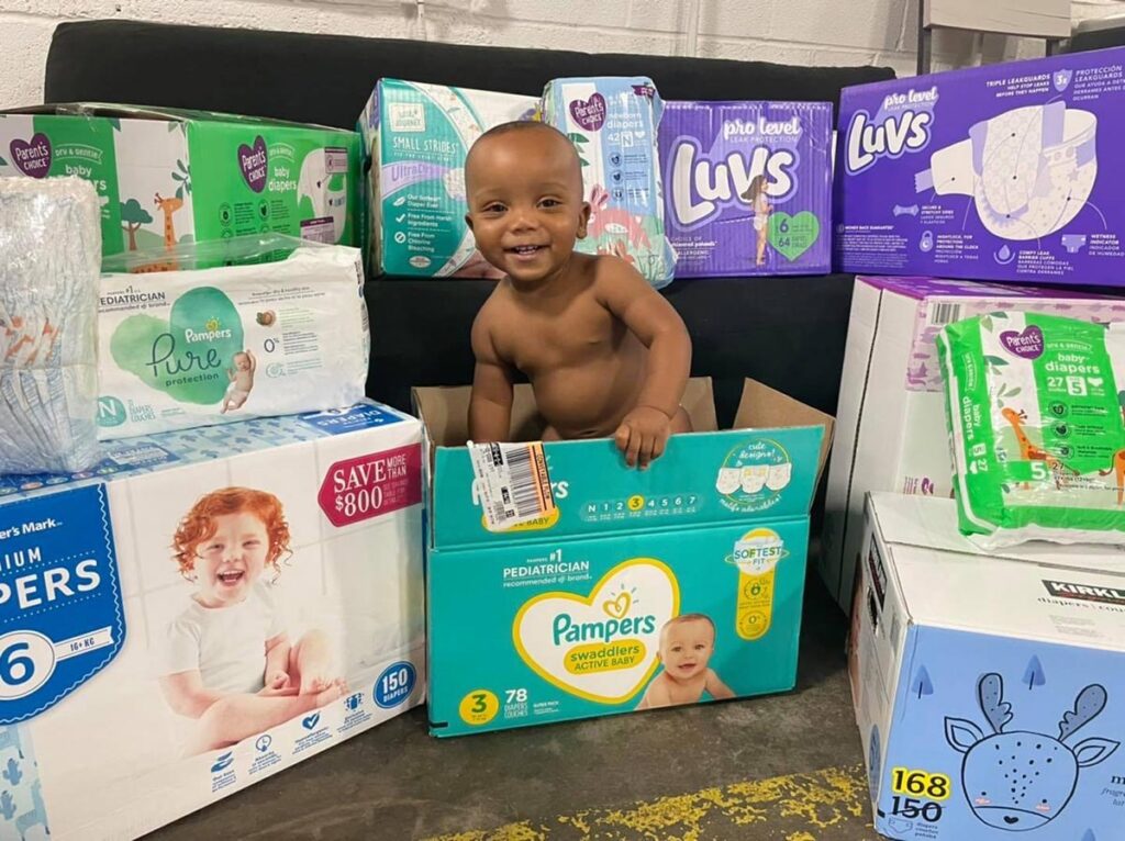 a cute baby peeks out of a box of diapers, surrounded by more boxes of diapers
