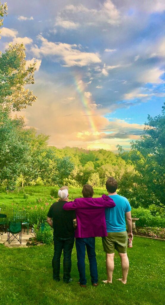 three people with their backs to the camera stand in a lush green landscape looking at a beautiful sky and rainbow