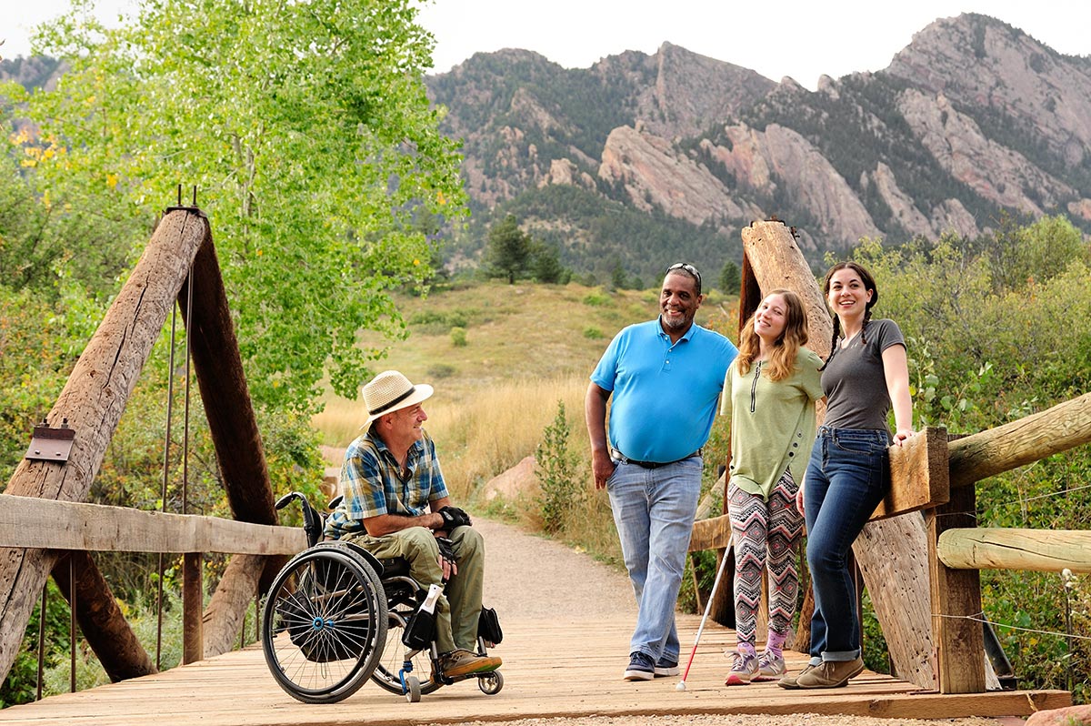 a man in a wheelchair next to three people standing on a bridge near beautiful mountains