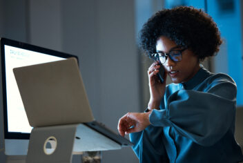 woman with glasses sitting in front of a laptop speaking on the phone
