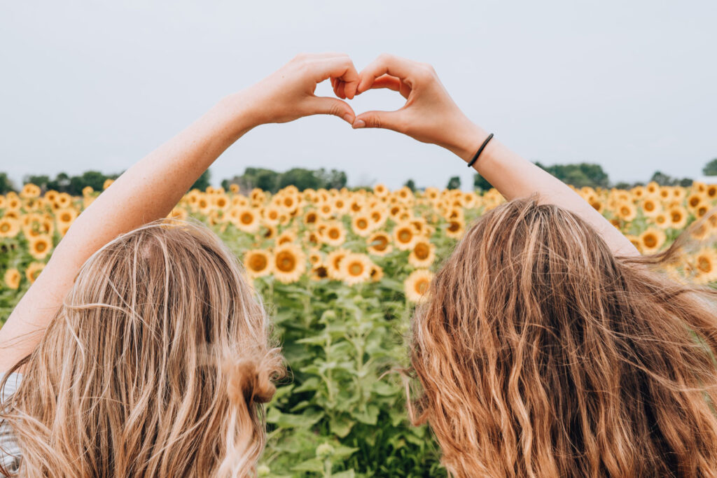 two girls in a field of sunflowers make a heart shape with their hands