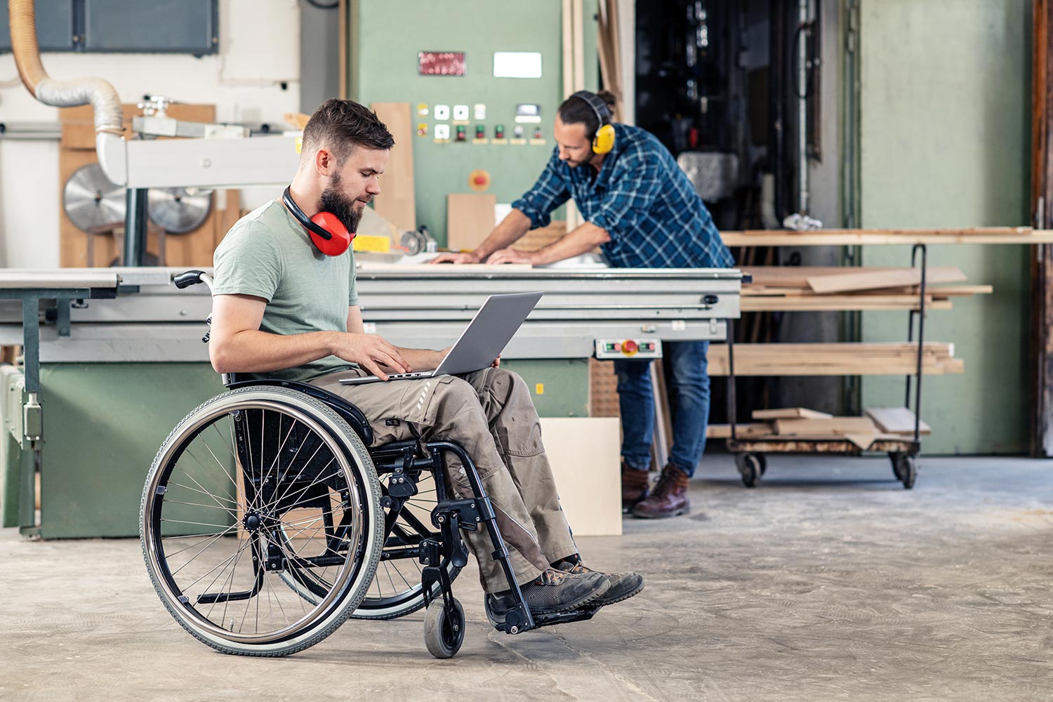 Man in wheelchair uses laptop, in the background a carpenter with boards at a table