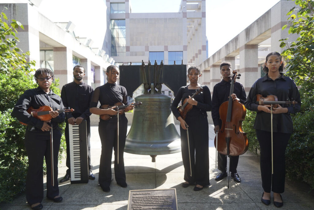 people with instruments stand around a large bell