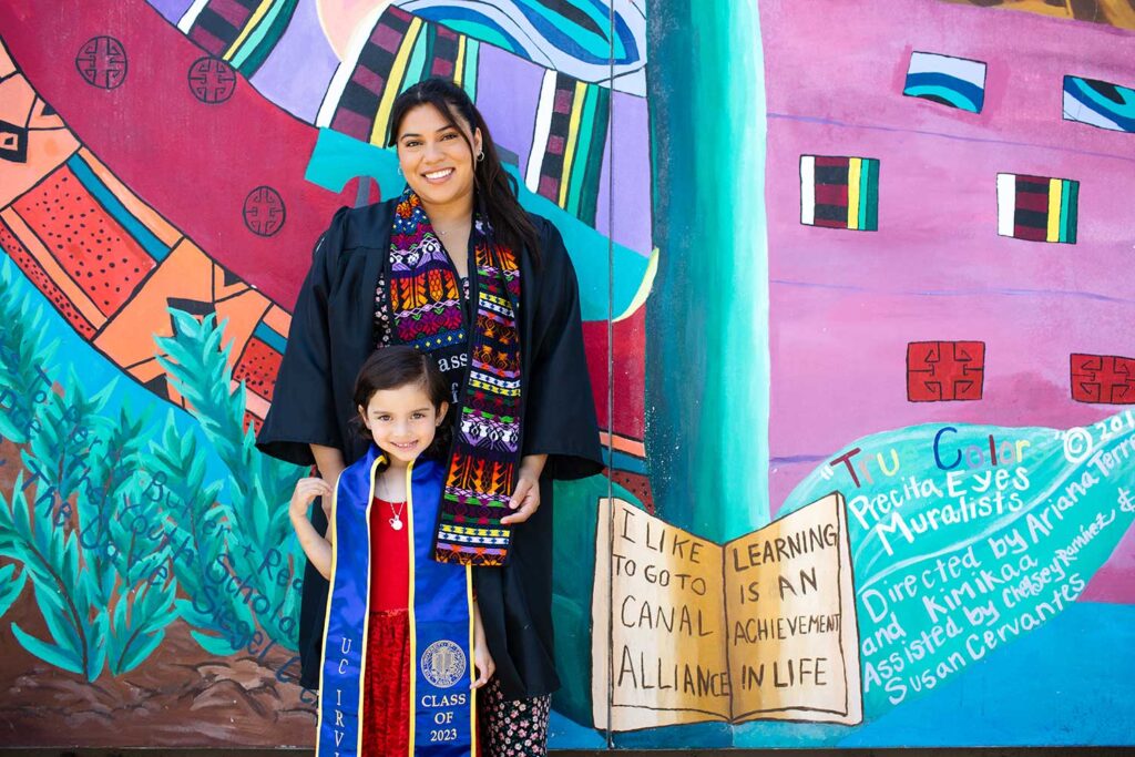 a woman in a graduation gown stands with a young boy in front of a school mural