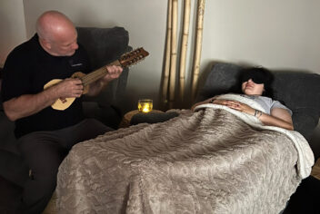 person playing a small guitar next to a young women resting in a bed