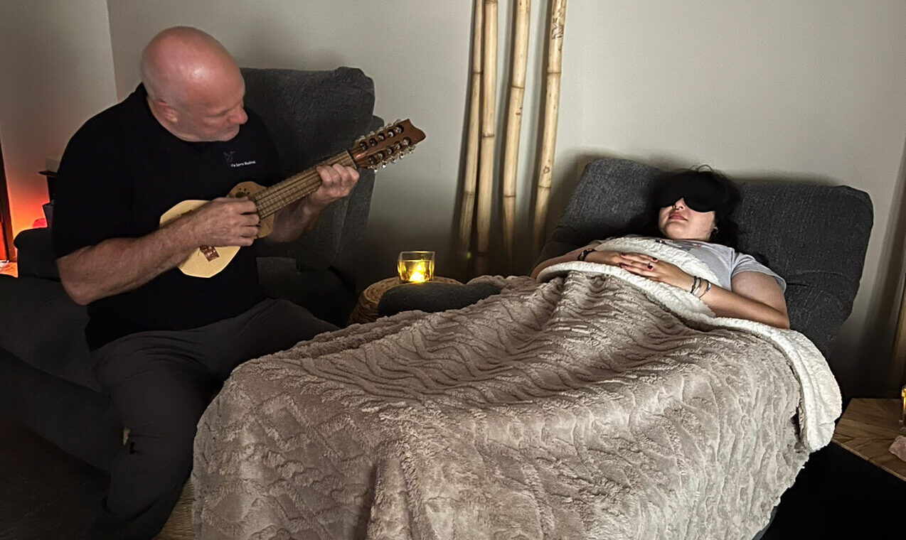 person playing a small guitar next to a young women resting in a bed