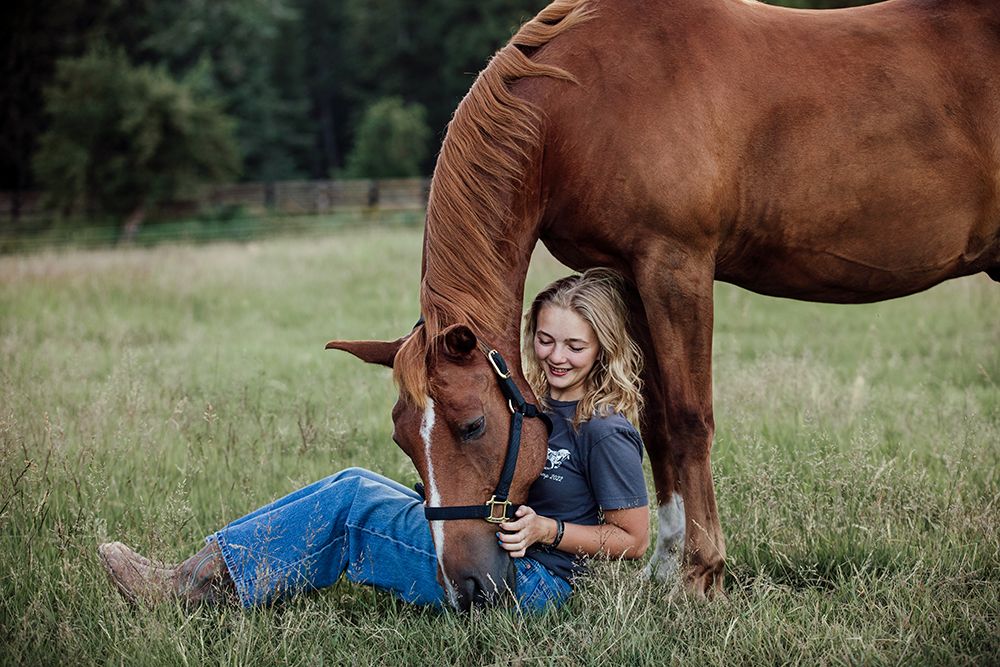 a young person sitting on the ground with a horse bending down next to them