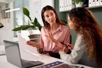 two women talking at a desk with a laptop