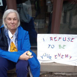 a woman sitting outside next to a sign that reads: I refuse to be your enemy