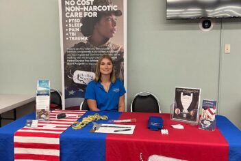 A woman sits at a display table, a sign in the back reads: No Cost Non-Narcotic Care for PTSD, Sleep, Pain, TBI, Trauma